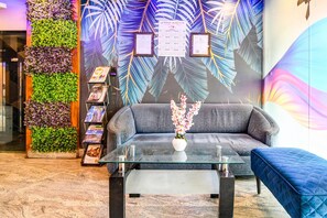 Un área de recepción con un sofá, una mesa de centro de vidrio, una estantería y una pared decorativa con motivos de hojas.