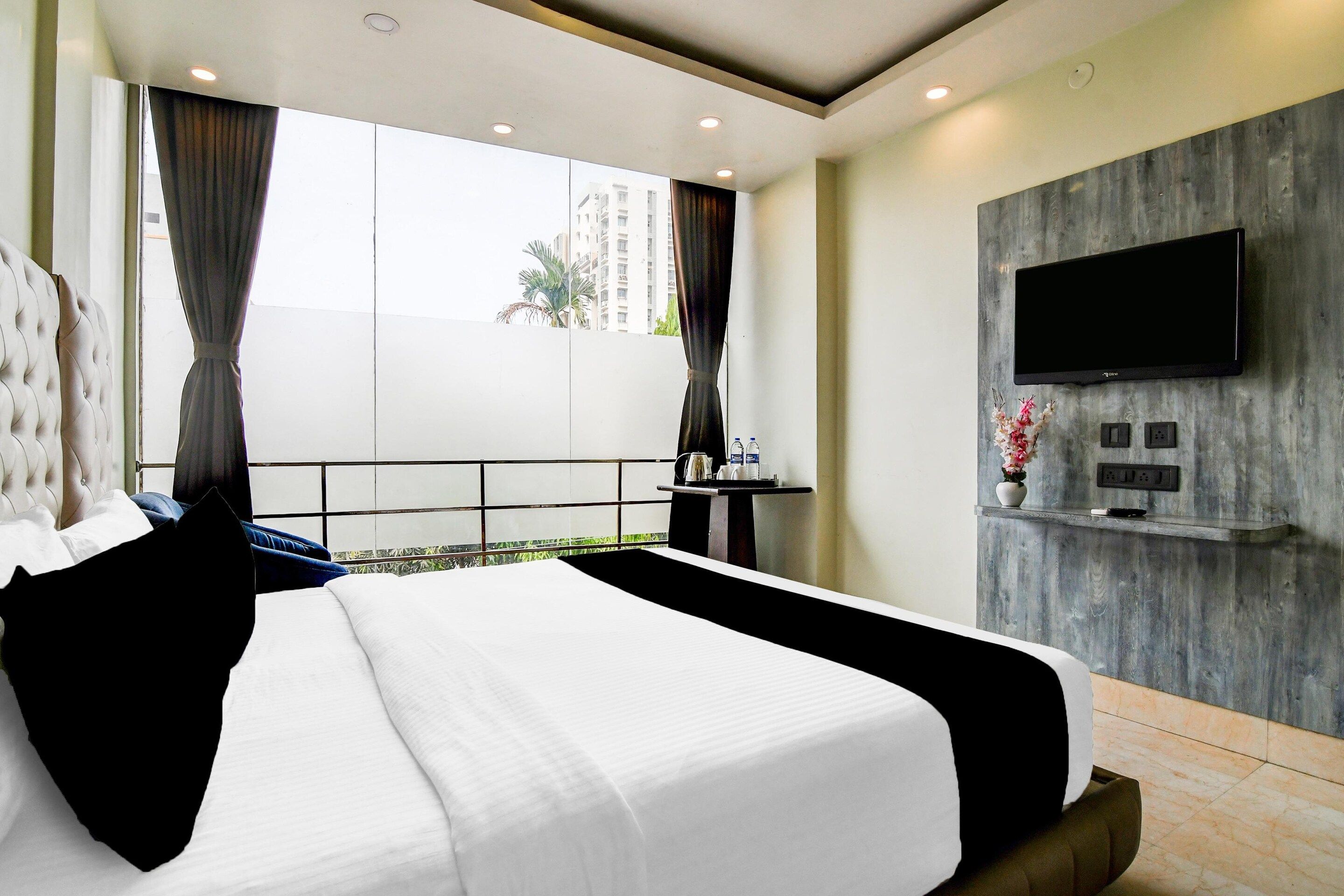 A hotel room with a large bed, a flat-screen TV mounted on a wooden wall, and a balcony with a view of buildings.