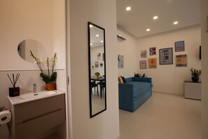 Una sala de estar moderna con un sofá azul, un comedor con mesa, y una pared blanca con obras enmarcadas.