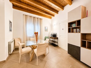 Ein modernes Wohnzimmer mit einem Glastisch, beigen Stühlen, einer Holzbalkendecke, einem Bücherregal und einer Heizung.
