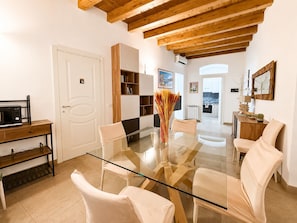 Una sala de estar moderna con una mesa de comedor de vidrio, sillas blancas, una estantería de madera y una cocina visible al fondo.