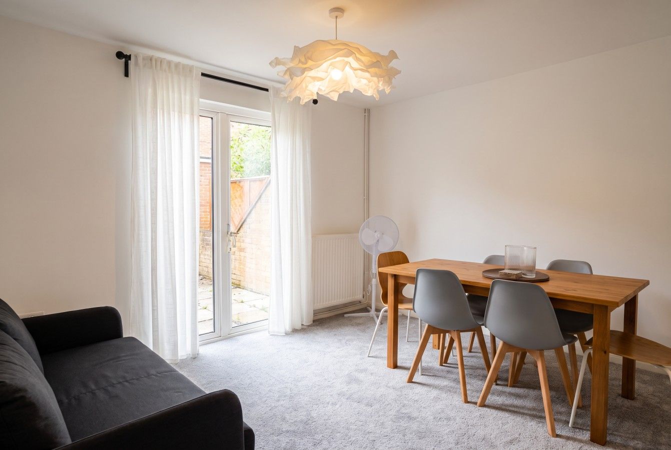 Een moderne woonkamer met een bank, een eettafel met stoelen, een plafondlamp en uitzicht door een glazen deur op een buitenruimte.