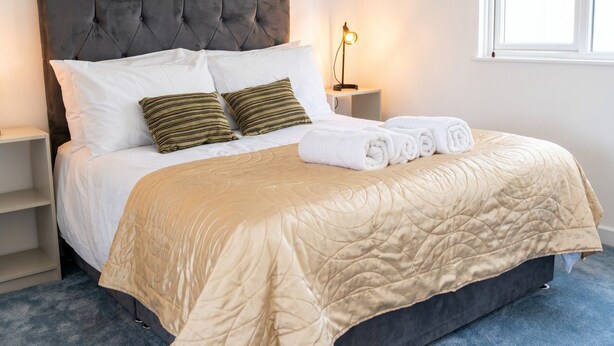 A neatly made bed with a tufted headboard, a bedside lamp, and a window with daylight. - Pet-Friendly 3-Bed House with Garden · Sleeps 6 in Headington, Oxford (Headington)