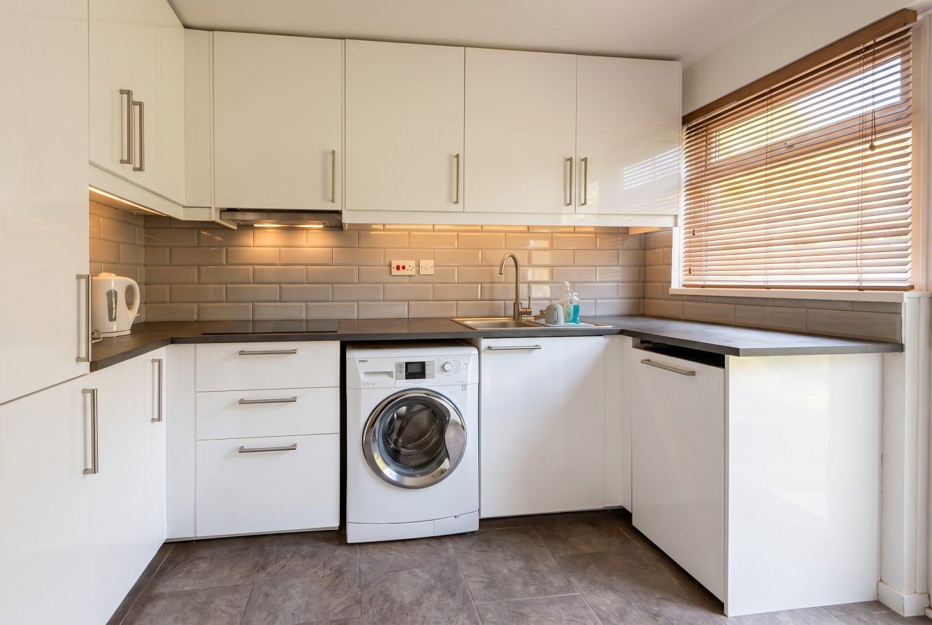 A compact kitchen with white cabinetry, a built-in washing machine, and a window with blinds.