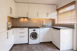 A compact kitchen with white cabinetry, a built-in washing machine, and a window with blinds.