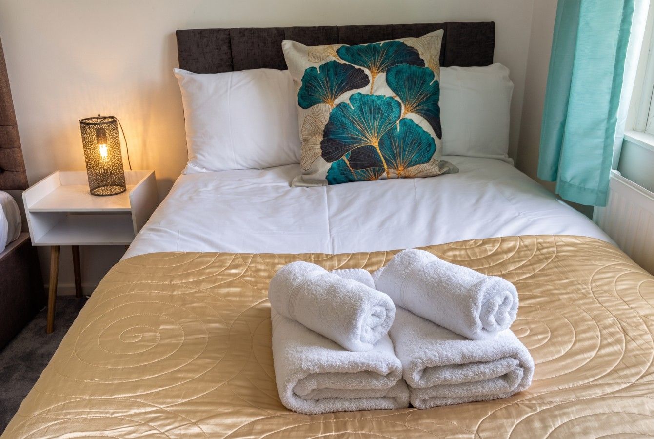 Un lit bien fait, recouvert d’une couvre-lit dorée, agrémenté d’un coussin décoratif et d’une pile de serviettes pliées.