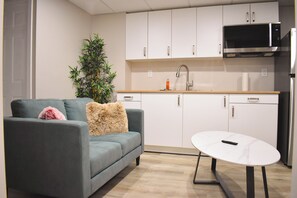 Un espacio habitable compacto con sofá, una mesita, una kitchenette con armarios y microondas, y una planta en maceta.