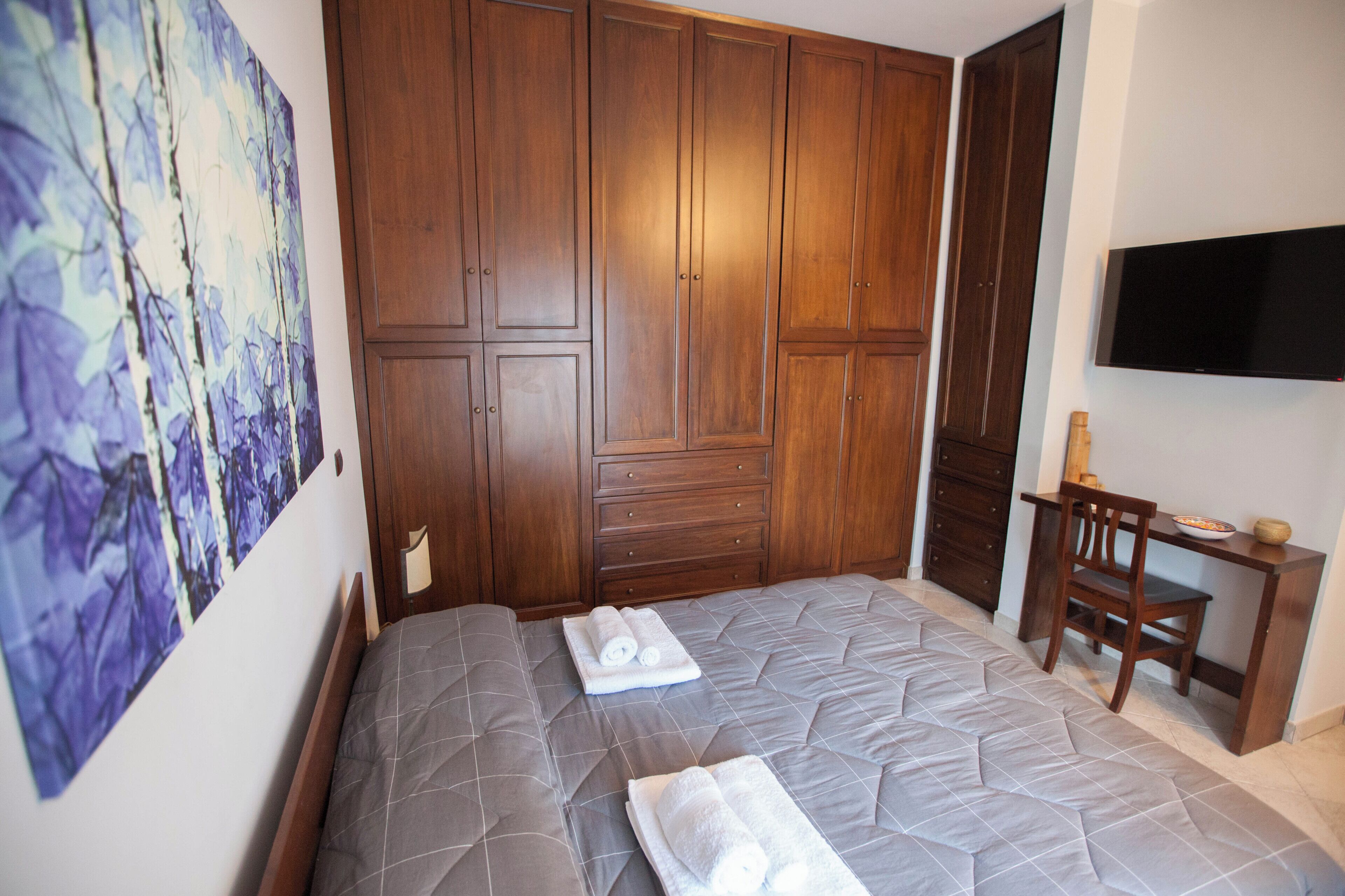 A bedroom with a large wardrobe, a bed with a quilted duvet, a desk with a chair, and a flat-screen TV mounted on the wall.