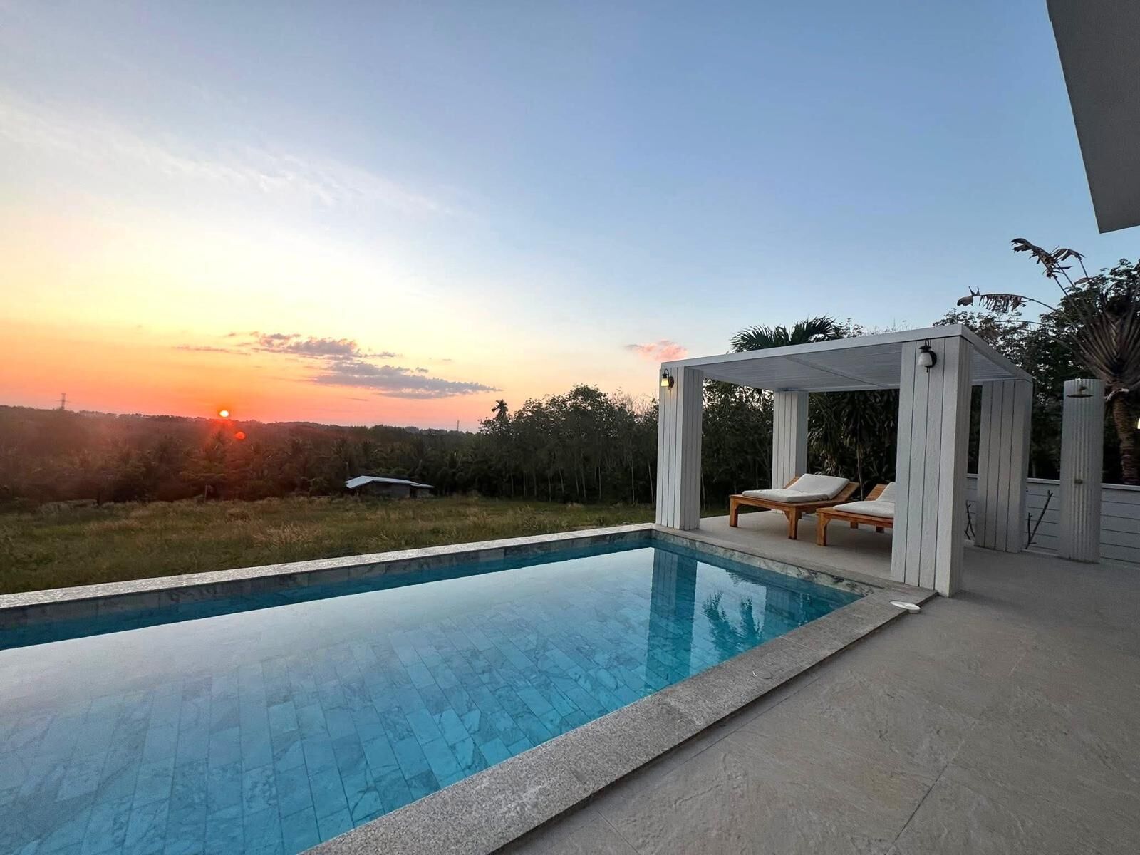 A swimming pool with a clear view of the sunset, a covered seating area with lounge chairs, and a natural landscape in the background.