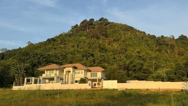 Um edifício residencial de dois andares com telhado plano, cercado por um gramado e uma colina com vegetação densa.