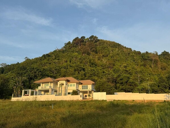 A two-story residential building with a flat roof, surrounded by a grassy area and a hill with dense vegetation. - Panoramic Sunset View Private Pool Villa, Maikhao beach (Thalang)