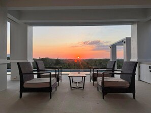 A balcony with four wicker chairs and a small table, overlooking a sunset with a view of the landscape.
