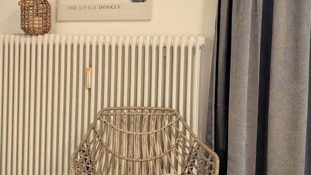 A room with a wicker chair, a radiator, a framed picture of a donkey, and a curtain. - Vrbo Property (Inzell)