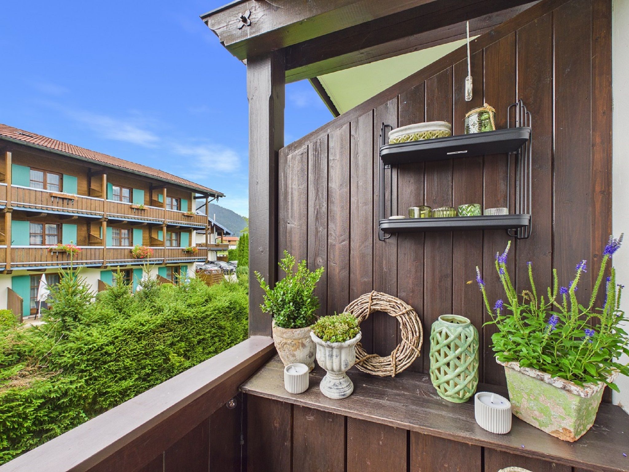 A wooden balcony with potted plants, a wicker wreath, and shelves with decorative items.