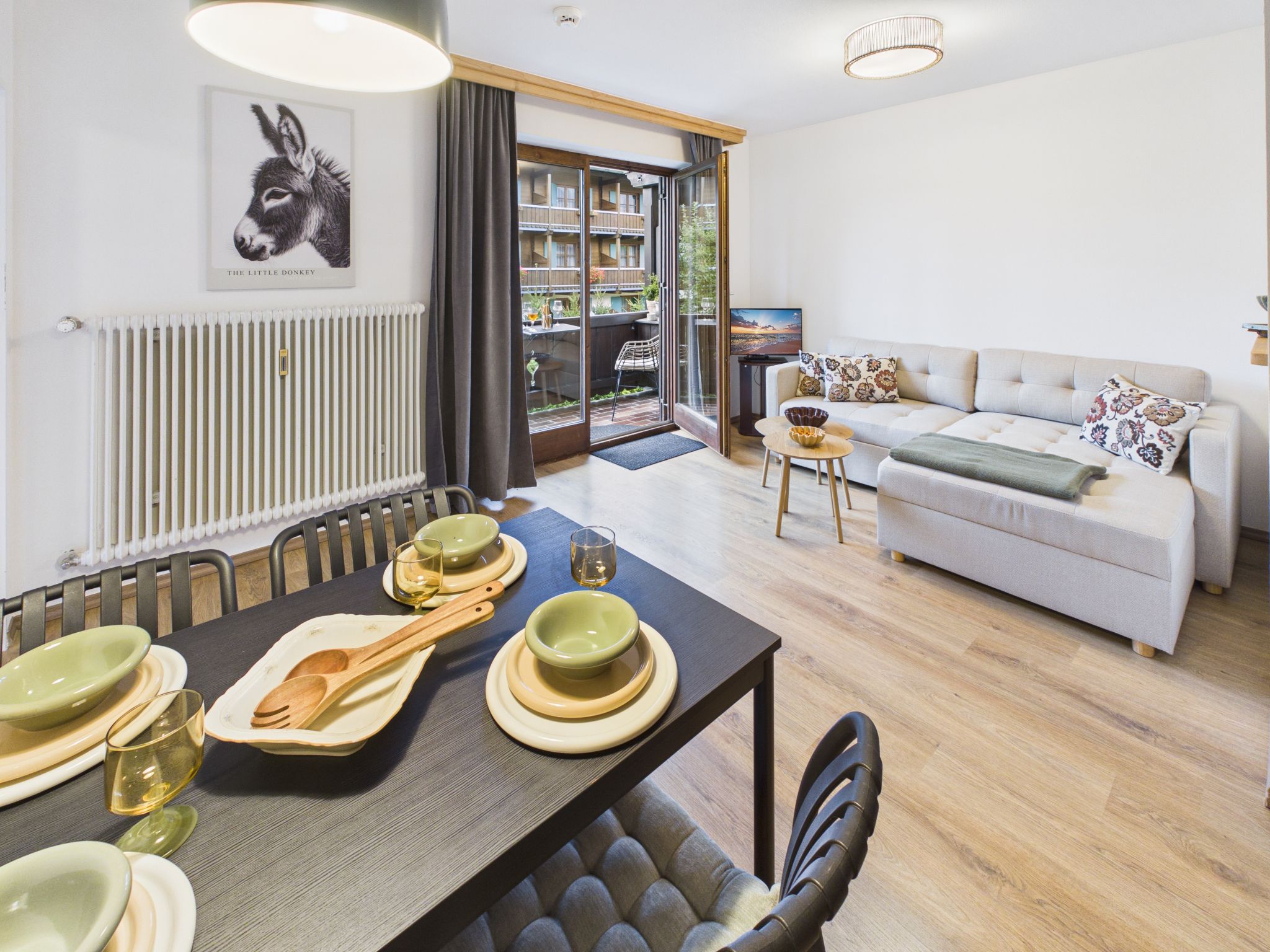 Een moderne woonkamer met een bank, een eettafel met borden en bestek, en een groot raam met uitzicht op een balkon.