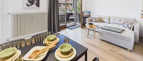 Een moderne woonkamer met een bank, een eettafel met borden en bestek, en een groot raam met uitzicht op een balkon.