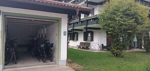 A building with a garage door, bicycles inside, and a grassy area with a bench and trees. - Vacation rental for 4 people, 1 separate bedroom, balcony, 54 m² (Inzell)