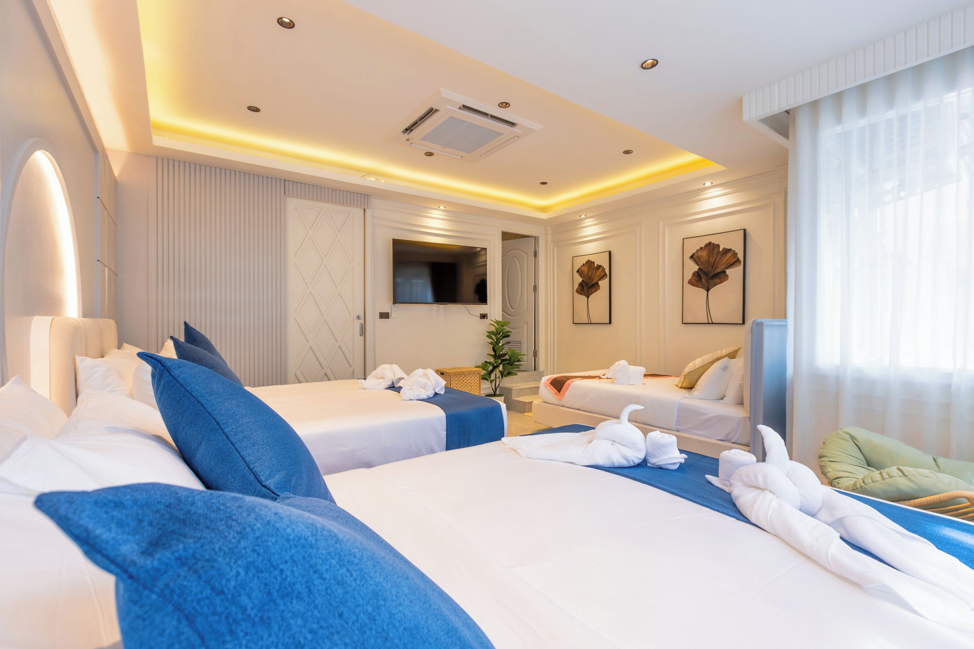 A hotel room with two beds, a flat-screen TV, artwork on the walls, and a large window with curtains.