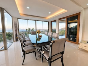 A modern dining room with a glass table and chairs, large windows offering a view of the landscape, and a balcony with a view. - Smith & Appy Seaview Penthouse (Koh Samui)