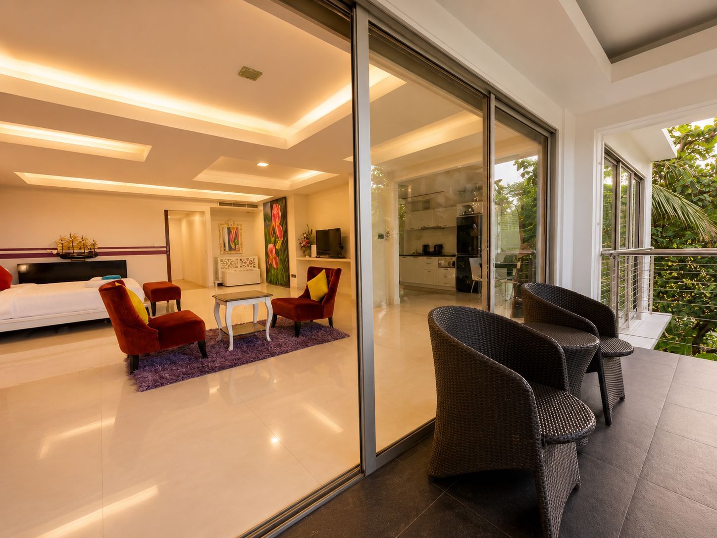 Una habitación de hotel moderna con una cama, dos sillas rojas, una mesa blanca, un televisor y un balcón con mobiliario exterior.