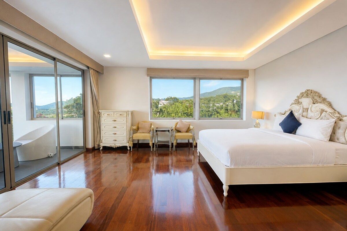 A spacious bedroom with a large bed, a chair, a small table, and a dresser. There are large windows offering a view of greenery and hills.