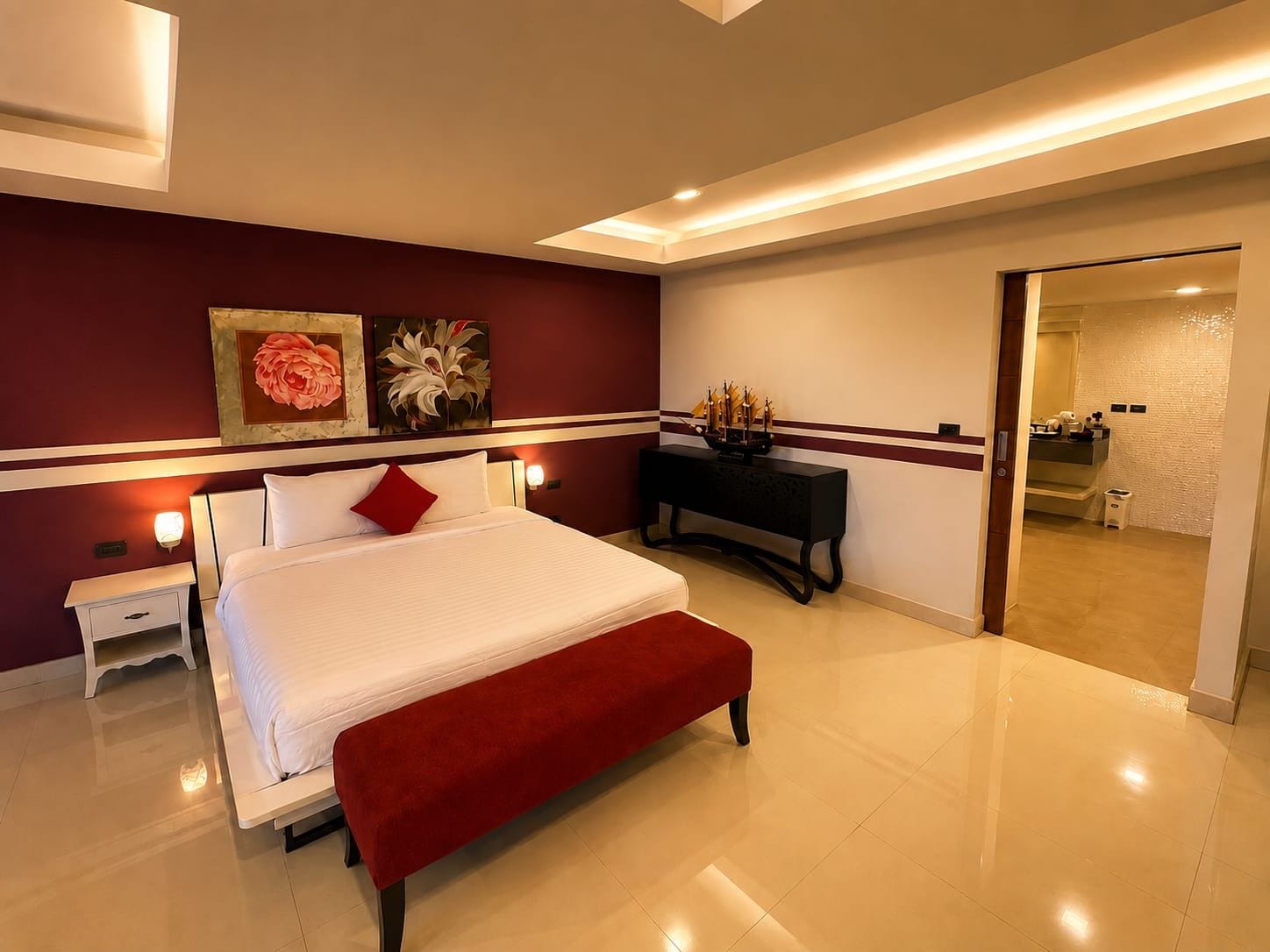 A modern hotel room with a large bed, a bedside table, a bench, a console table with decorative items, and a view into the bathroom.