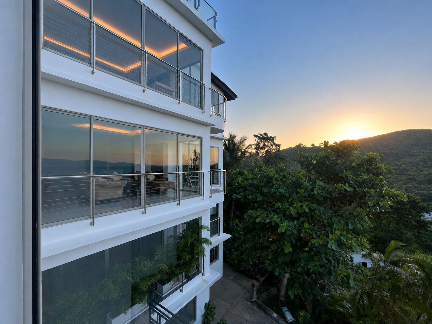 A modern multi-story building with large windows, a balcony, and a view of a sunset over a hilly landscape.