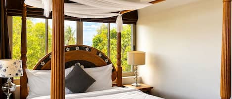 A four-poster bed with a canopy, a bedside table with a lamp, and a view of greenery outside.