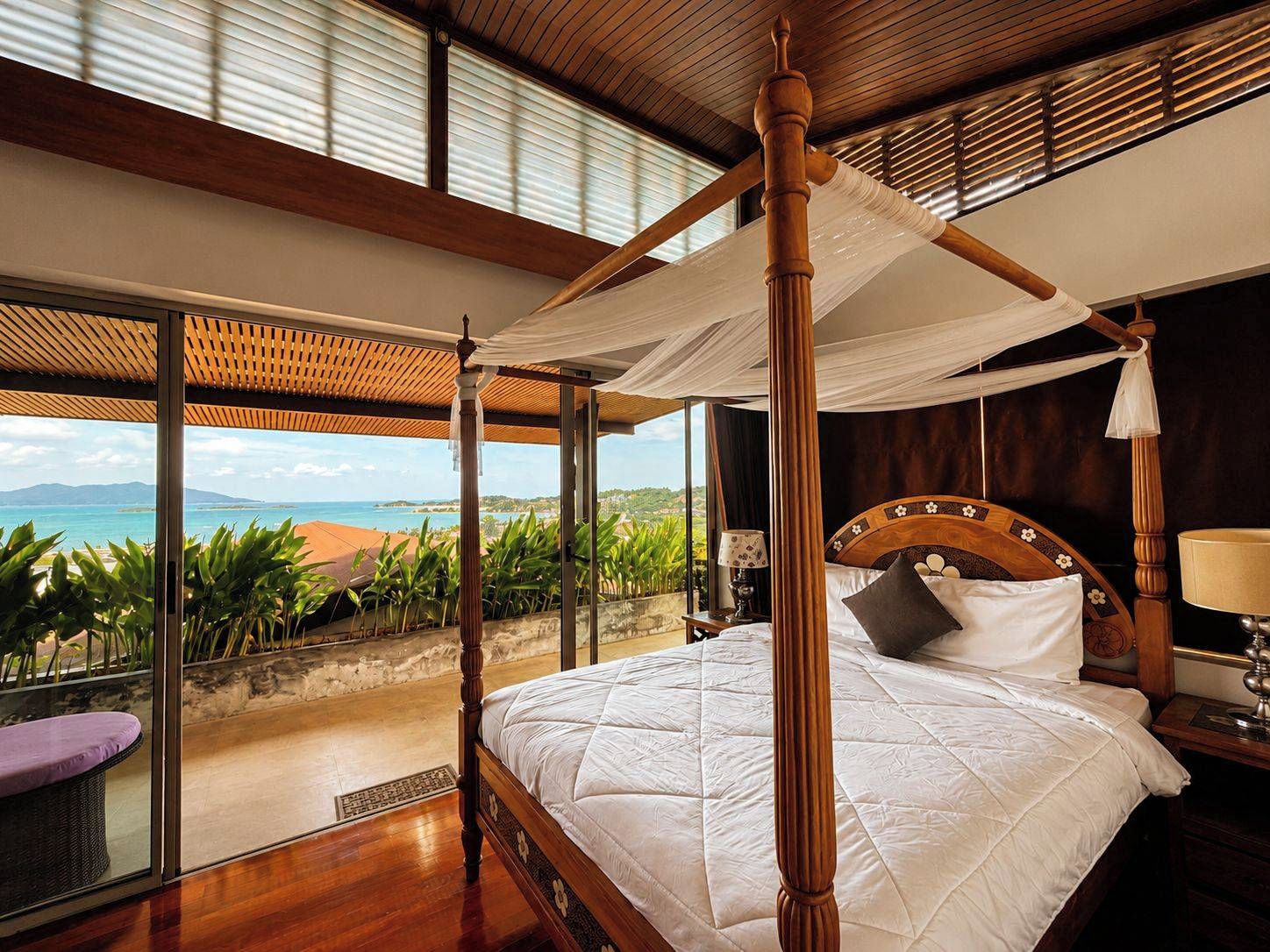A four-poster bed with a canopy, a wooden headboard, and a view of the sea through large windows.