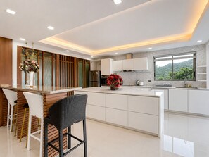 A modern kitchen with a central island, white cabinetry, and a view of greenery outside. - Smith & Appy Seaview Penthouse (Koh Samui)