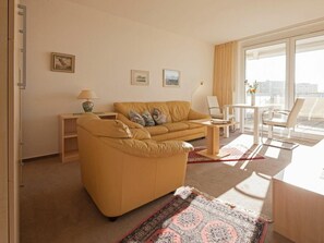A living room with a sofa, a coffee table, and a dining area with chairs. There are framed pictures on the wall and a window with curtains. - Rotter/seelord 27 (Norderney)