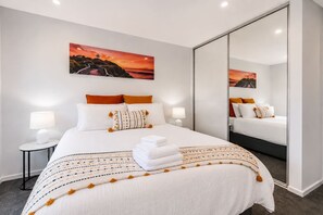 Ein modernes Hotelzimmer mit einem großen Bett, zwei Nachttischen, einem Schminktisch, einem großen Spiegel und einem Gemälde eines Sonnenuntergangs.