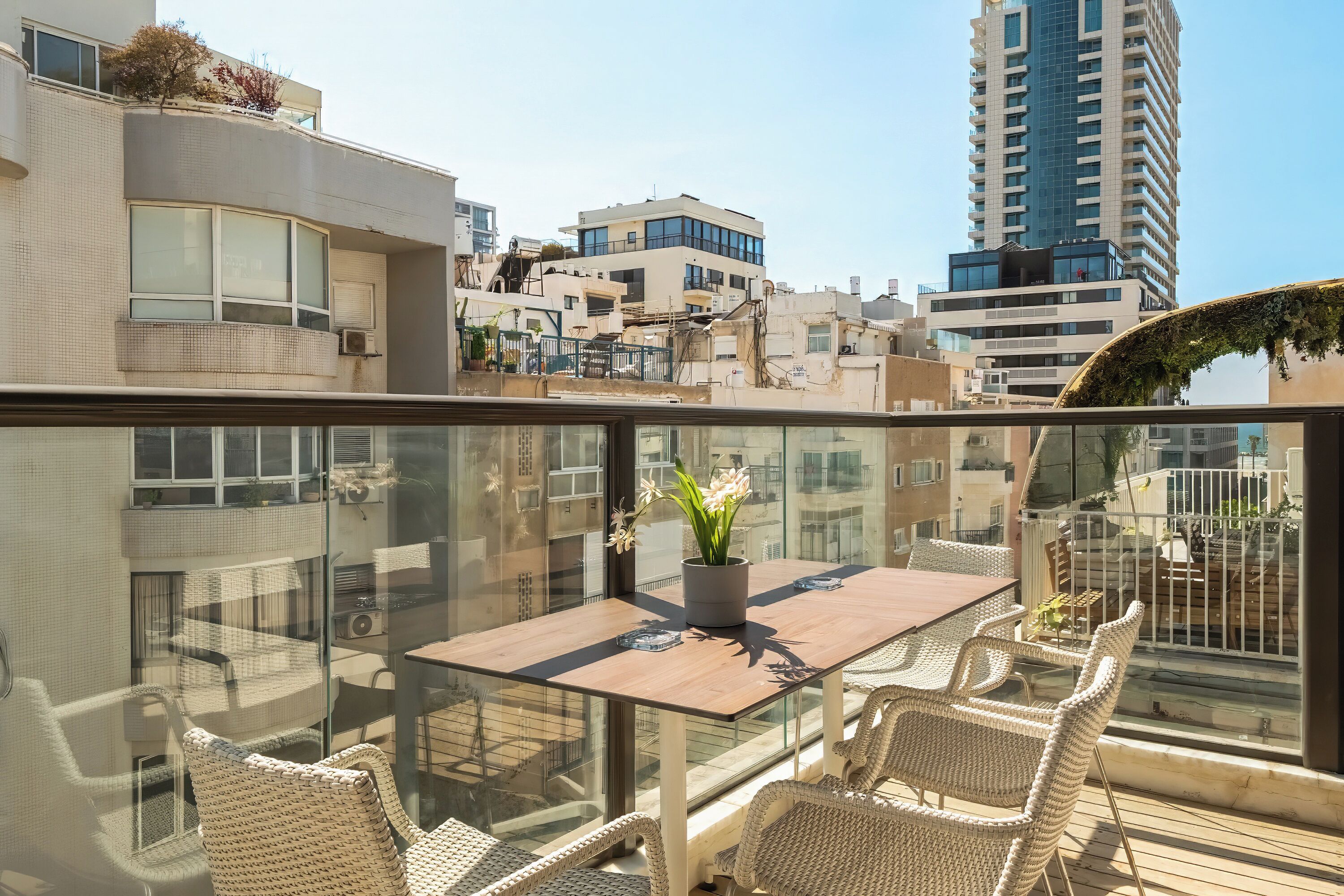 A rooftop terrace with a glass balustrade, a wooden table, wicker chairs, and a potted plant.