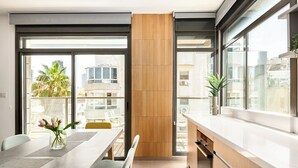 A modern kitchen with a large window, a dining area with a table and chairs, and a view of the cityscape. - Sea Breeze – Luxury 3BR Beachfront Apartment on Zrubavel Street (Tel Aviv-Yafo)