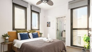A modern bedroom with a large bed, two windows, a ceiling fan, and a bathroom visible through a glass door. - Sea Breeze – Luxury 3BR Beachfront Apartment on Zrubavel Street (Tel Aviv-Yafo)