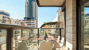 A balcony with a glass table and wicker chairs, overlooking a cityscape with modern buildings. - Sea Breeze – Luxury 3BR Beachfront Apartment on Zrubavel Street (Tel Aviv-Yafo)