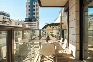 Un balcon doté d’une table en verre et de chaises en osier, offrant une vue sur un paysage urbain avec des bâtiments modernes.