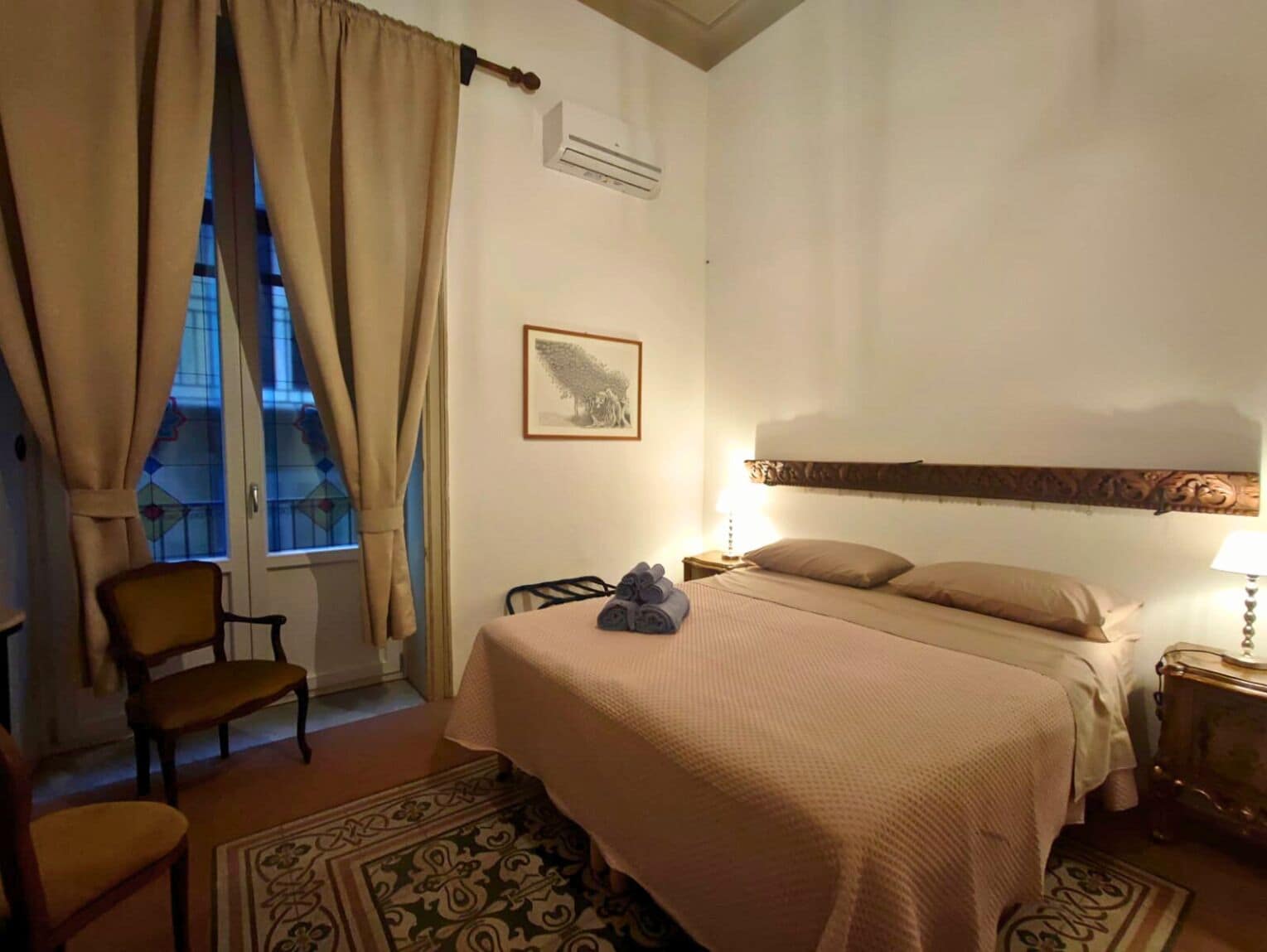 Quarto de hotel com cama, uma cadeira, mesa de cabeceira com luminária, janela com cortinas e um quadro na parede.