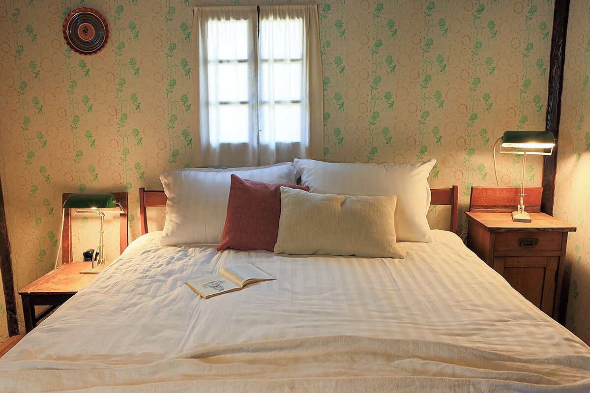 A neatly made bed with pillows and a book, a window with curtains, and a bedside lamp.
