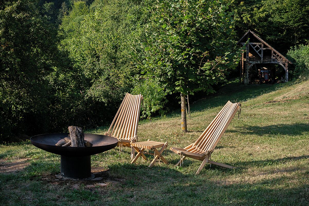 A grassy outdoor area with a fire pit, two wooden chairs, and a rustic wooden structure in the background.