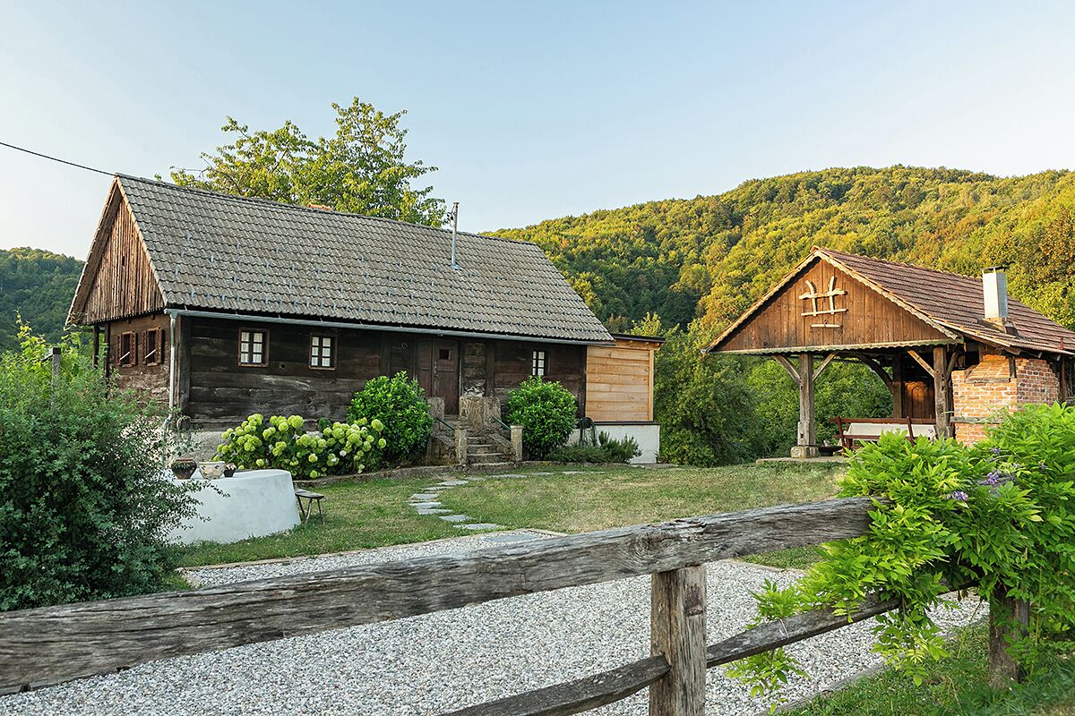 A rustic wooden house with a sloped roof and a small porch, surrounded by greenery and a gravel path.
