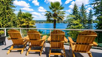 A deck with four wooden Adirondack chairs facing a body of water, surrounded by trees and a clear sky.