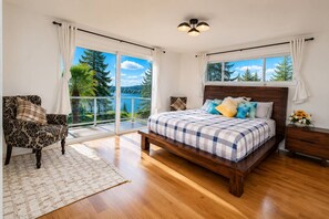 A bedroom with a wooden bed, a chair, a nightstand, and a view of the outdoors.