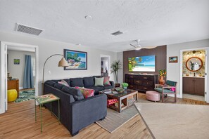 A living room with a dark sofa, a coffee table, and a TV. There is a plant, a lamp, and a painting on the wall. - Hibiscus Hideaway – Tropical Waterfront Paradise Villa 26 (Cape Coral)