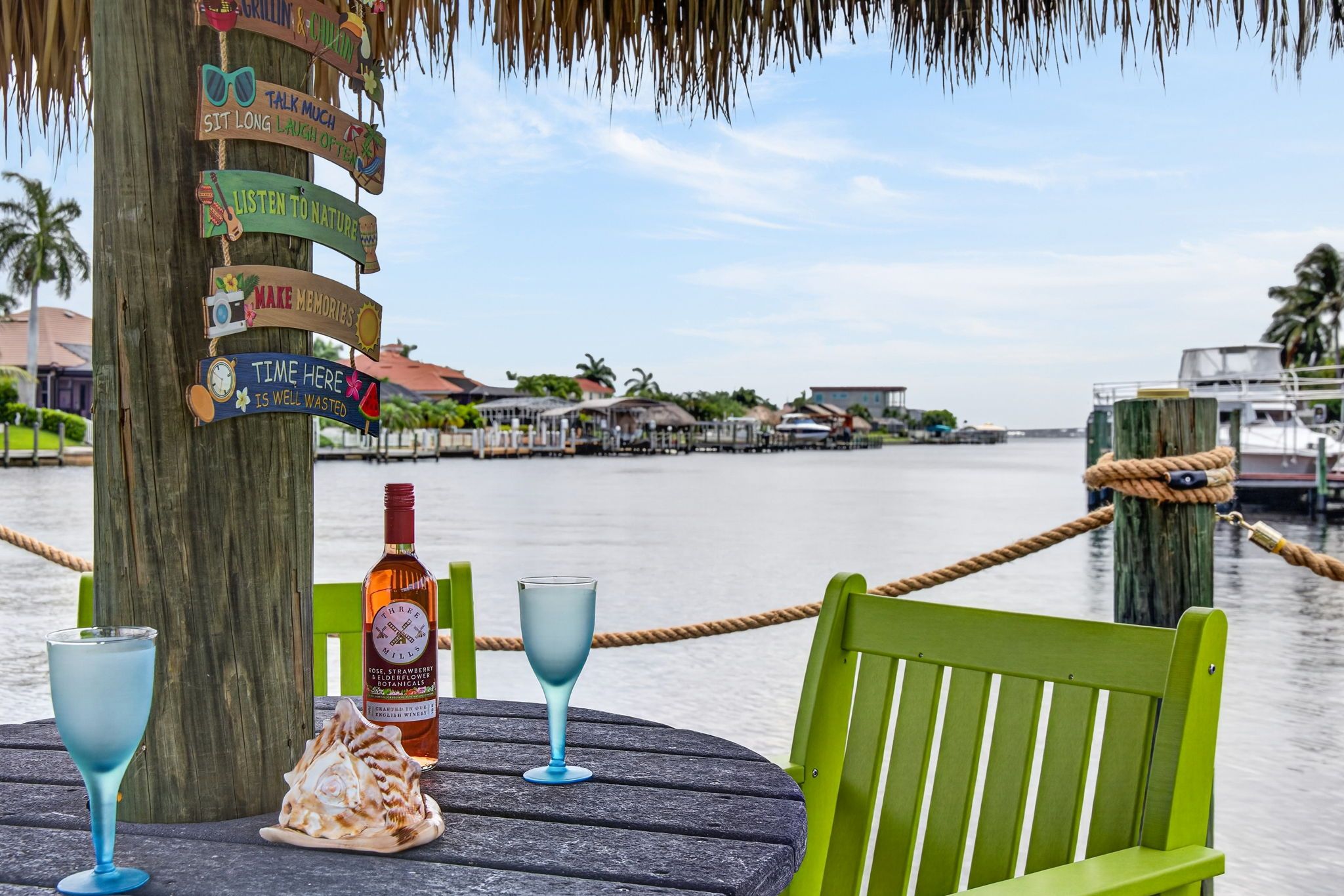A wooden signpost with various signs, a bottle of Three Mills Rose, Strawberry & Elderflower Botanical, two glasses, and a seashell on a wooden table by a waterfront.