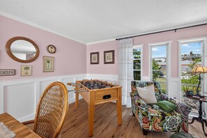 A cozy room with a foosball table, a wicker chair, a colorful sofa, and a view through large windows. - Hibiscus Hideaway – Tropical Waterfront Paradise Villa 26 (Cape Coral)