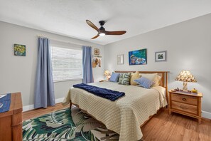 A bedroom with a bed, a nightstand, a lamp, a ceiling fan, and a window with curtains. - Hibiscus Hideaway – Tropical Waterfront Paradise Villa 26 (Cape Coral)