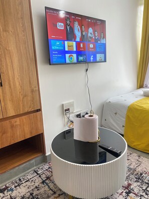 Ein Hotelzimmer mit einem an der Wand befestigten Fernseher, der verschiedene Streaming-Dienste anzeigt, einem runden Tisch mit einer Fernbedienung und einem Bett mit einer gelben Decke.