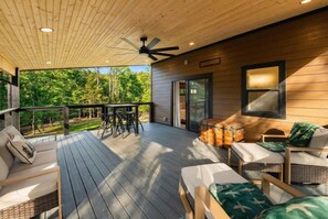 A covered outdoor deck with seating, a ceiling fan, and a view of the surrounding forest.