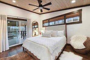 A bedroom with a large bed, a ceiling fan, a sliding glass door to a balcony, and a view of the outdoors.